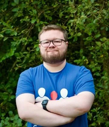 Andrew Greenslade, smiling with arms folded in front of green foliage, wearing a blue CoderDojo t-shirt.