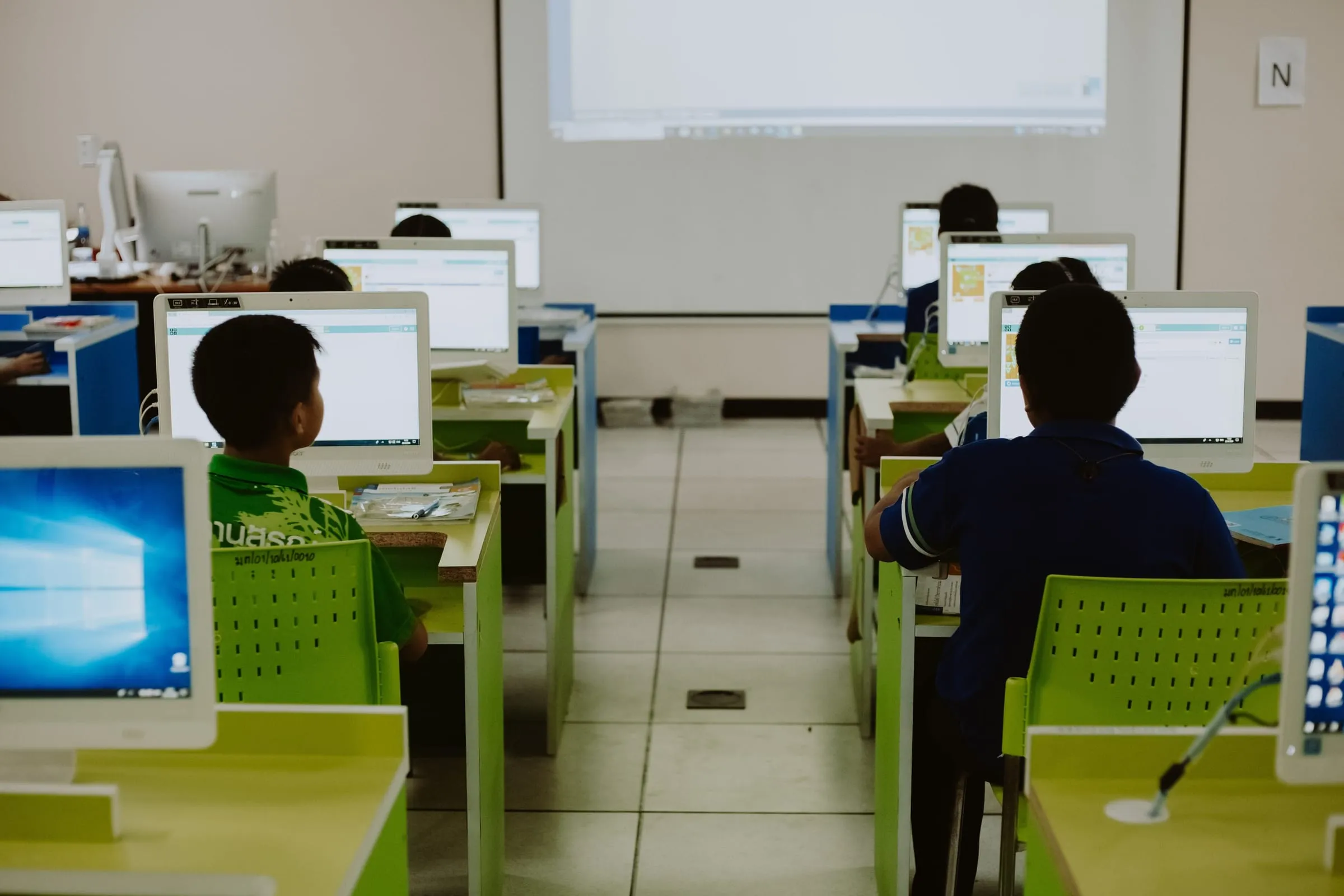 Young people at rows of computers in a classroom, seen from behind, facing a projector screen.