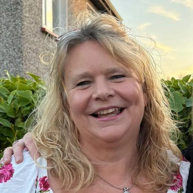 Susie Clince, smiling outdoors in a floral blouse with green hedges and a sunset sky behind her.
